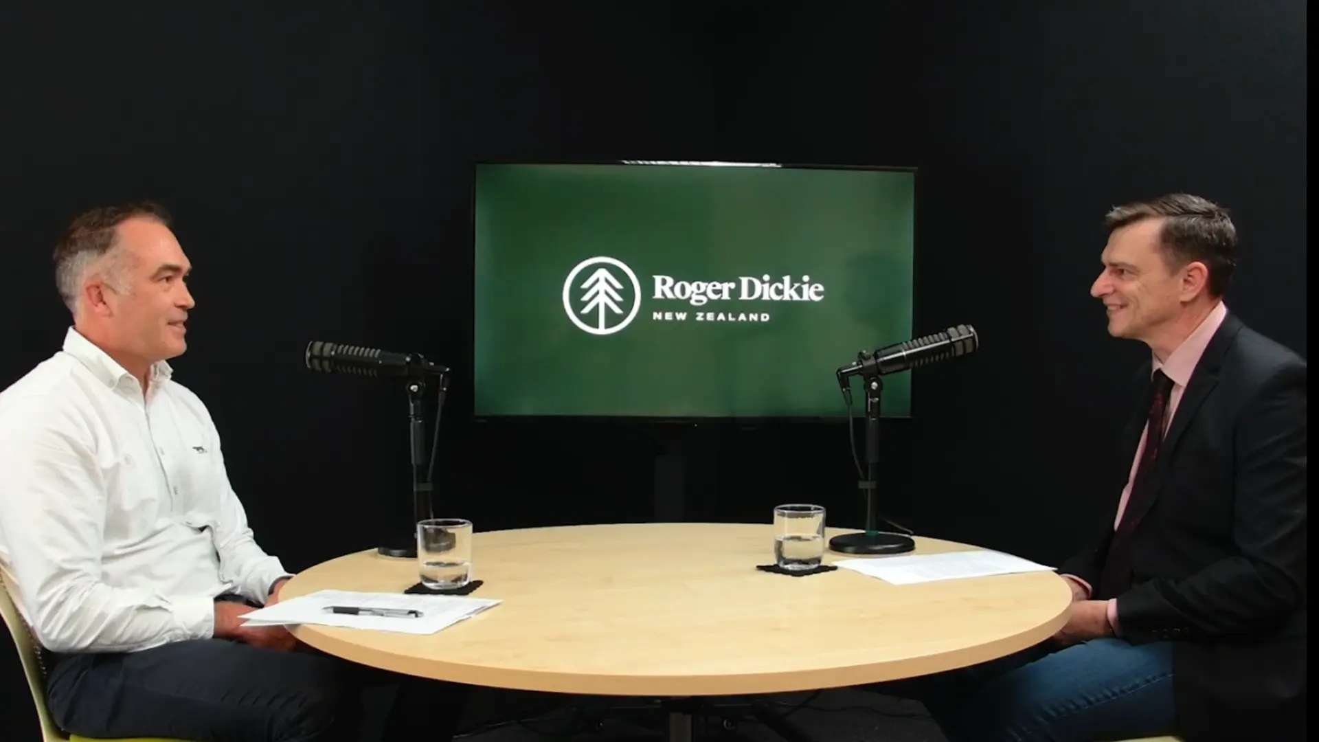 Two men sitting across from each other at a round table with microphones and Roger Dickie New Zealand logo behind.