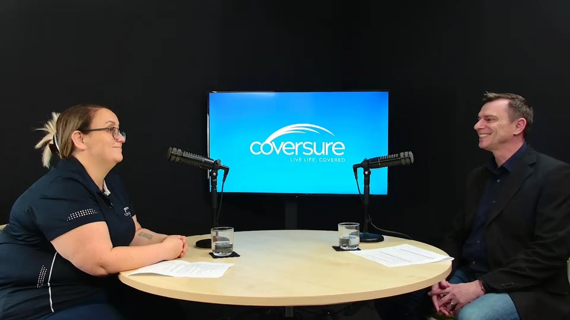 Two people seated at a round table with microphones and a screen showing the Coversure logo behind them.