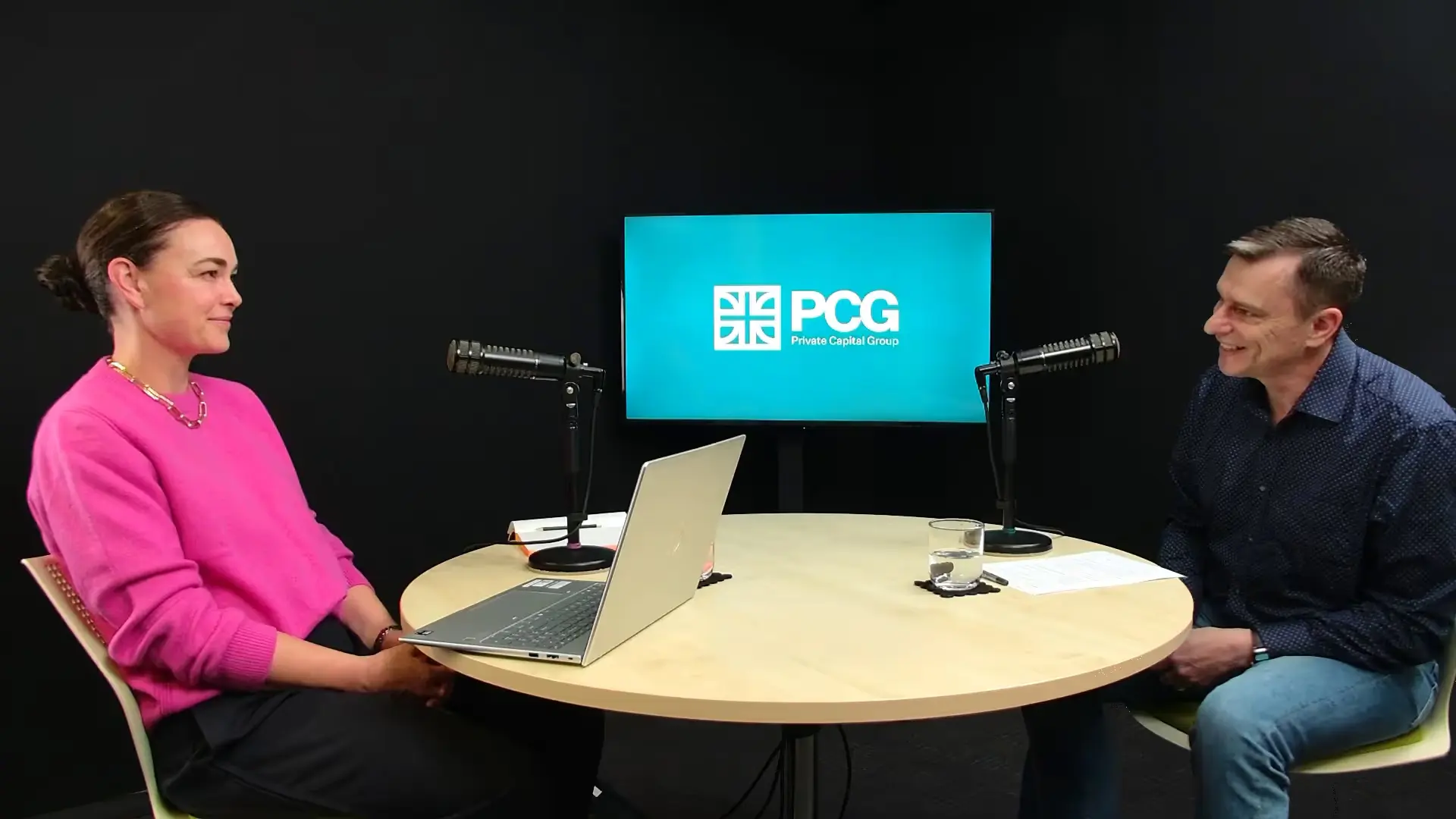 Two people sit facing each other at a round table with microphones and a laptop in a podcast studio.
