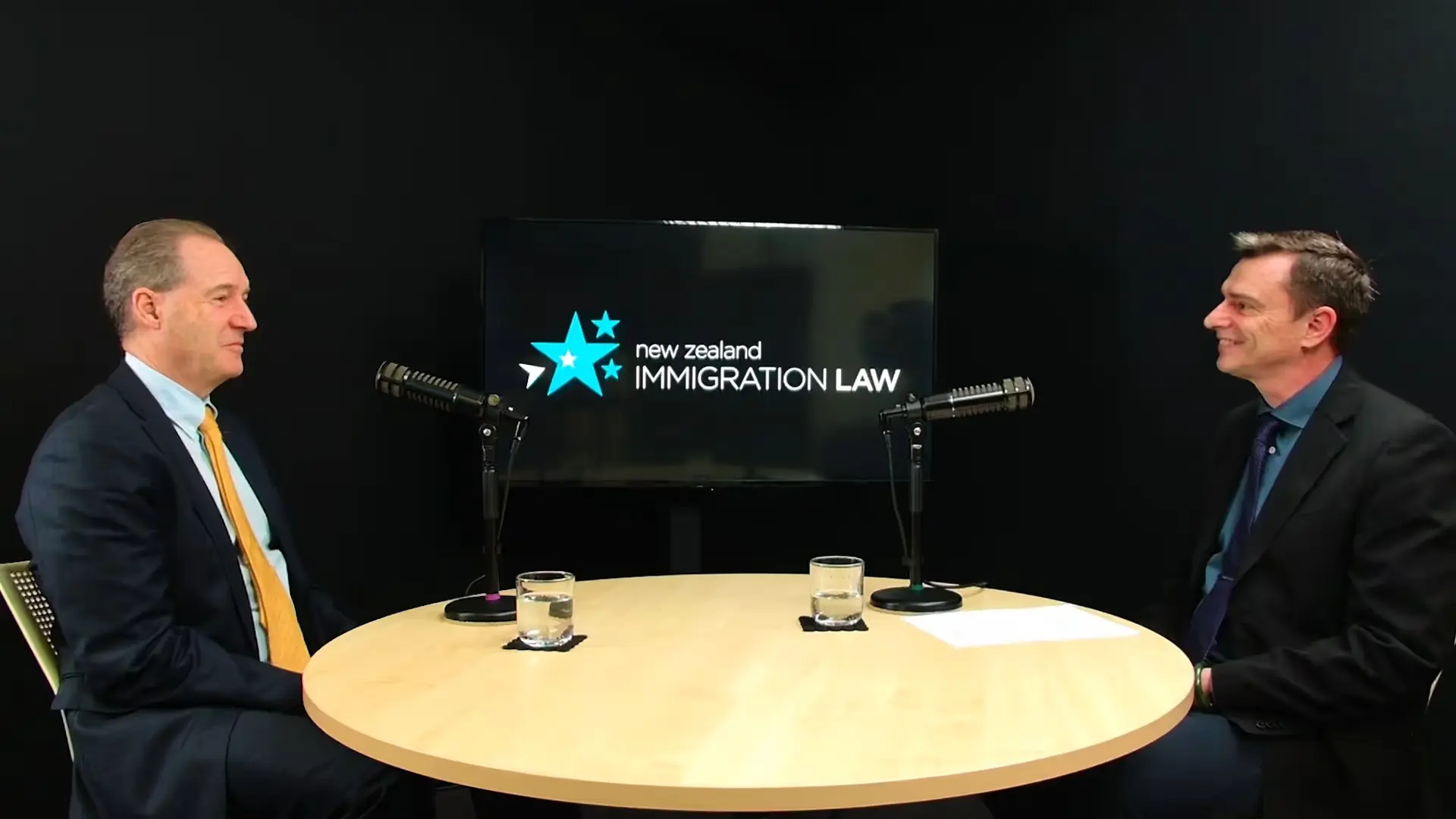 Two men in suits seated at a round table with microphones, New Zealand Immigration Law displayed on screen.