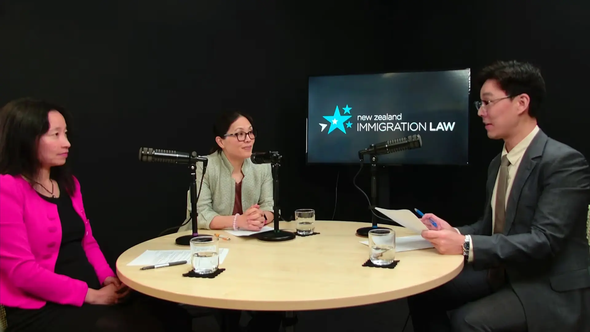 Three people seated around a table with microphones, discussing New Zealand immigration law.