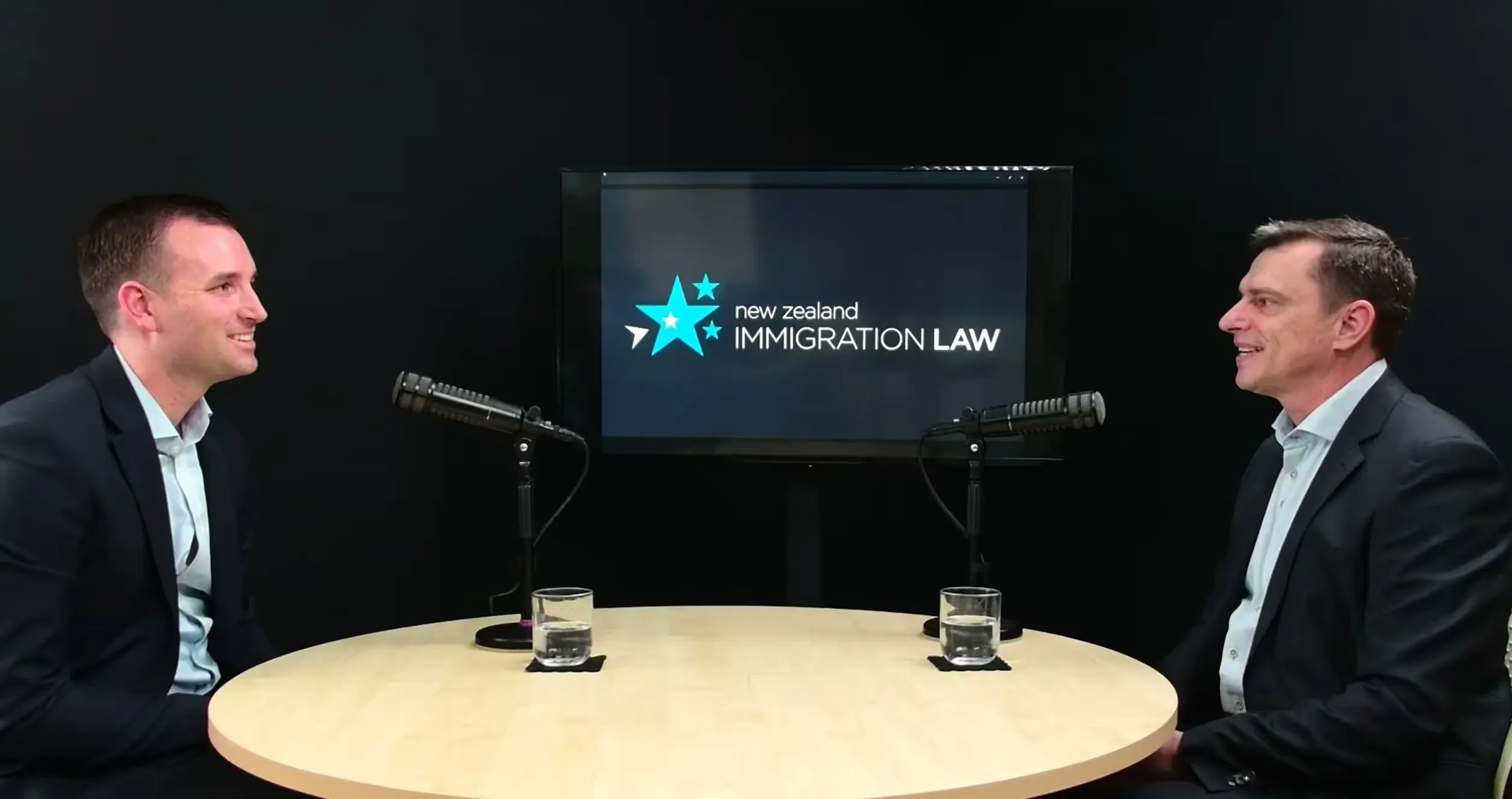 Two men in suits sit at a round table with microphones and water glasses, discussing New Zealand immigration law.