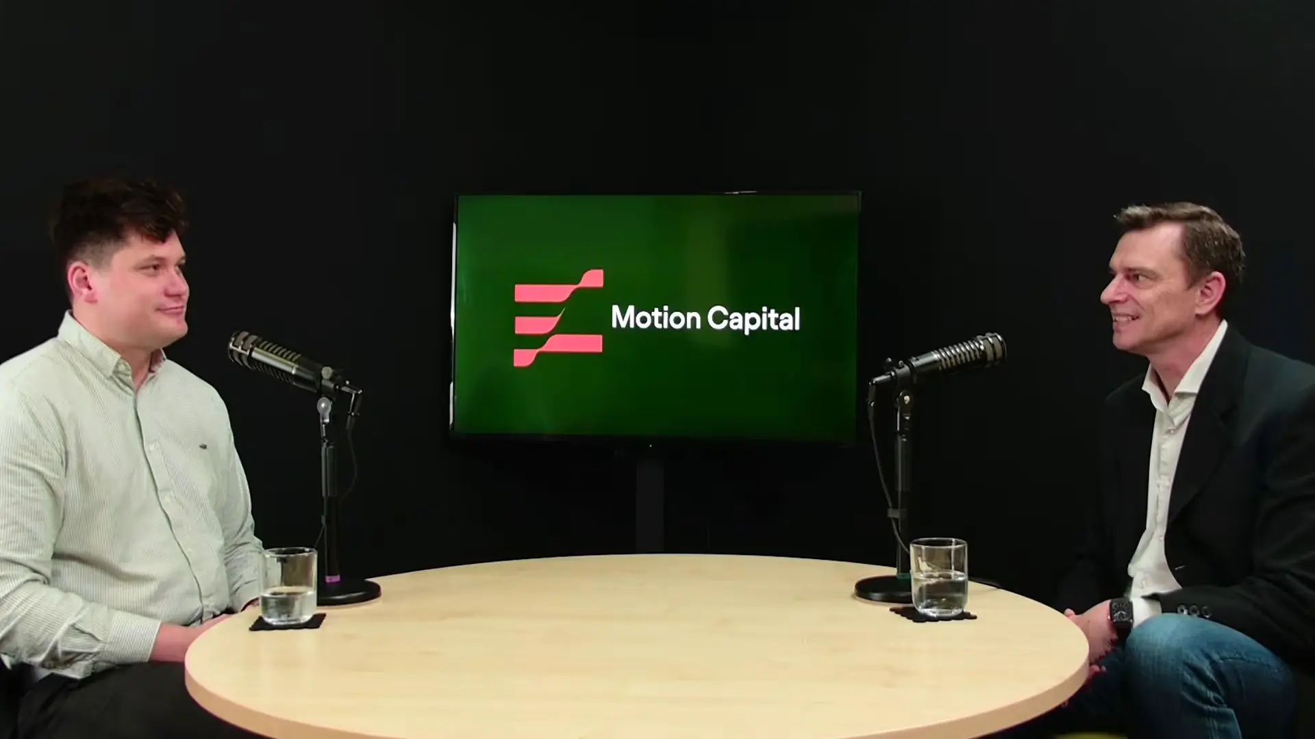 Two men seated at a round table with microphones, a screen behind shows the logo and text 'Motion Capital'.