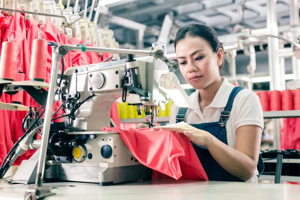 A garment worker sewing fabric at an industrial machine inside a clothing factory, representing labor conditions in the fashion industry.
