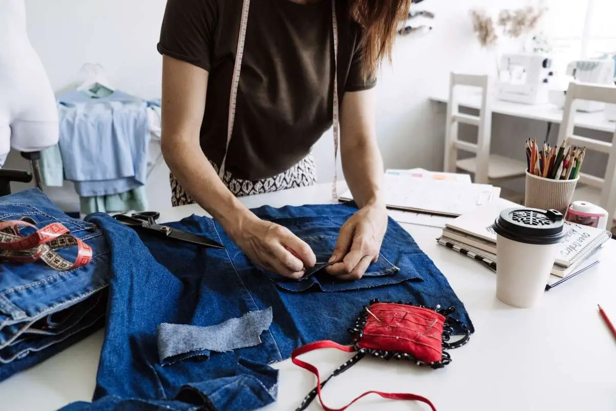 Hands repairing or upcycling denim fabric on a table with sewing tools, showing a sustainable approach to extending clothing life.