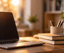 A closed laptop on a wooden desk with two stacked books and a white container holding pens, with a warmly lit blurred background.