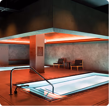 Indoor pool area with recessed lighting casting a warm orange glow, a stainless steel handrail leading into a lit rectangular pool, and two wooden chairs with cushions against a textured wall.