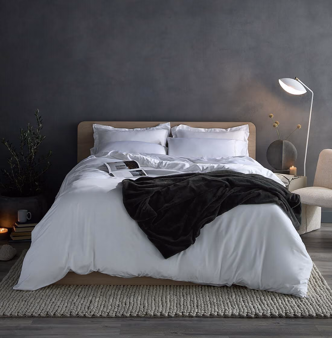 Modern bedroom with a neatly made bed featuring white bedding, four white pillows, and a dark gray blanket draped across the center. An open book lies on the bed. The bed sits on a textured light gray area rug on wood flooring. To the right, there is a beige upholstered chair, a white standing lamp, and a nightstand with a round gray vase holding dried flowers and a lit candle. The back wall is dark gray, creating a calm atmosphere. To the left, a small stack of books, a gray cup, a lit candle, and a plant in a dark pot complete the setting.