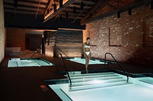 A woman in a bikini standing on steps leading into an illuminated indoor pool area with multiple pools and exposed brick walls.