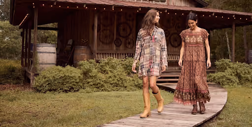 Two women walking on a wooden pathway in front of a rustic wooden cabin with barrels and greenery, both wearing patterned dresses and boots.
