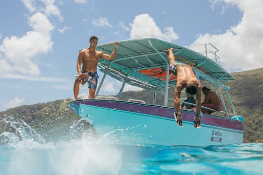 Two young men on a turquoise boat with purple trim in clear blue water under a partly cloudy sky; one man stands on the bow while the other dives off the boat into the water.