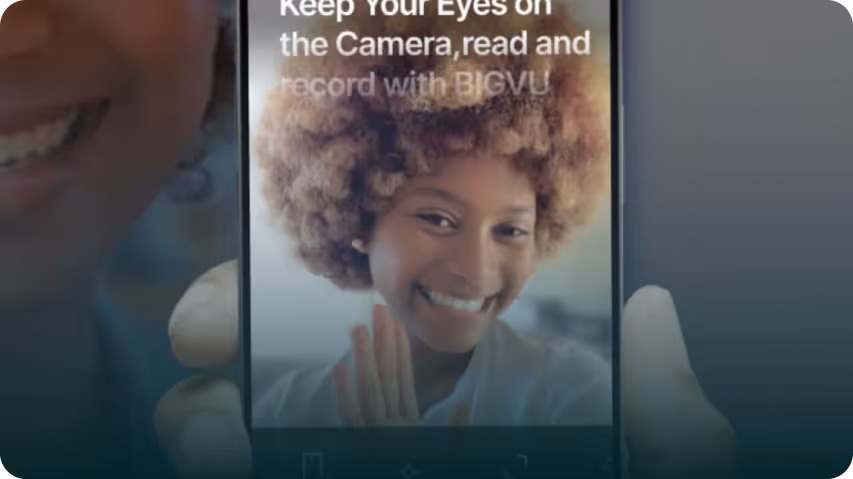 Smartphone screen showing a smiling woman with curly hair holding her hands together in front of her, with overlay text about keeping eyes on the camera and using BIGVU.