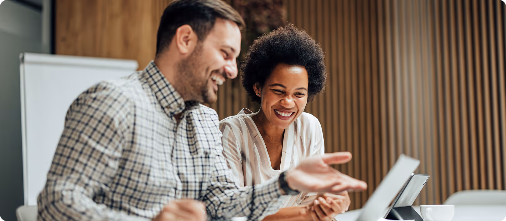Two coworkers smiling and having a lively discussion while looking at a laptop in an office.