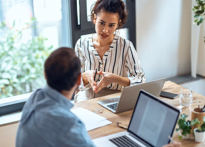 A woman in a striped blouse talking and gesturing with her hands during a meeting with a man at a wooden table with laptops and documents.