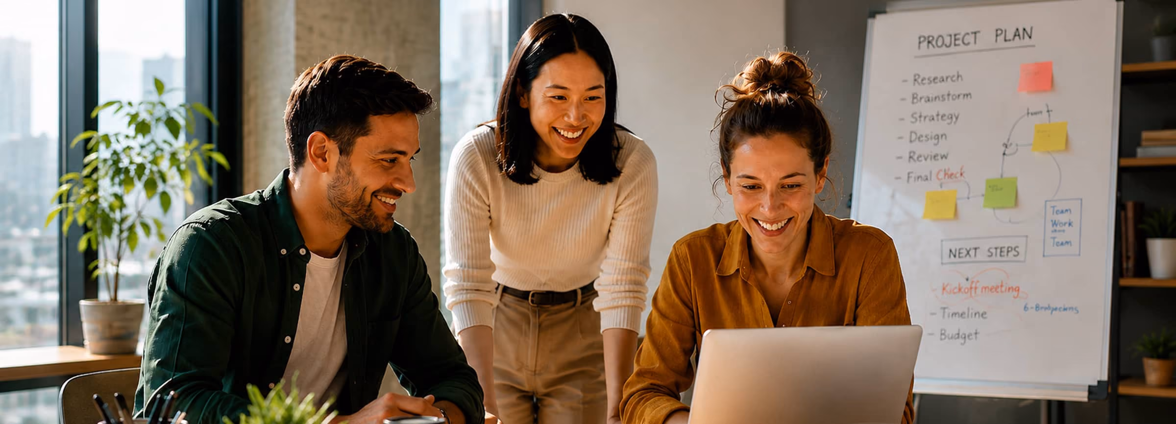 Three colleagues smiling and working together on a laptop in an office with a project plan board.