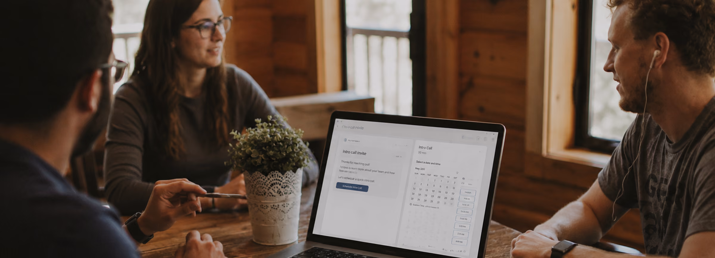 Three people sitting at a wooden table with a laptop open to a scheduling app and a small plant.