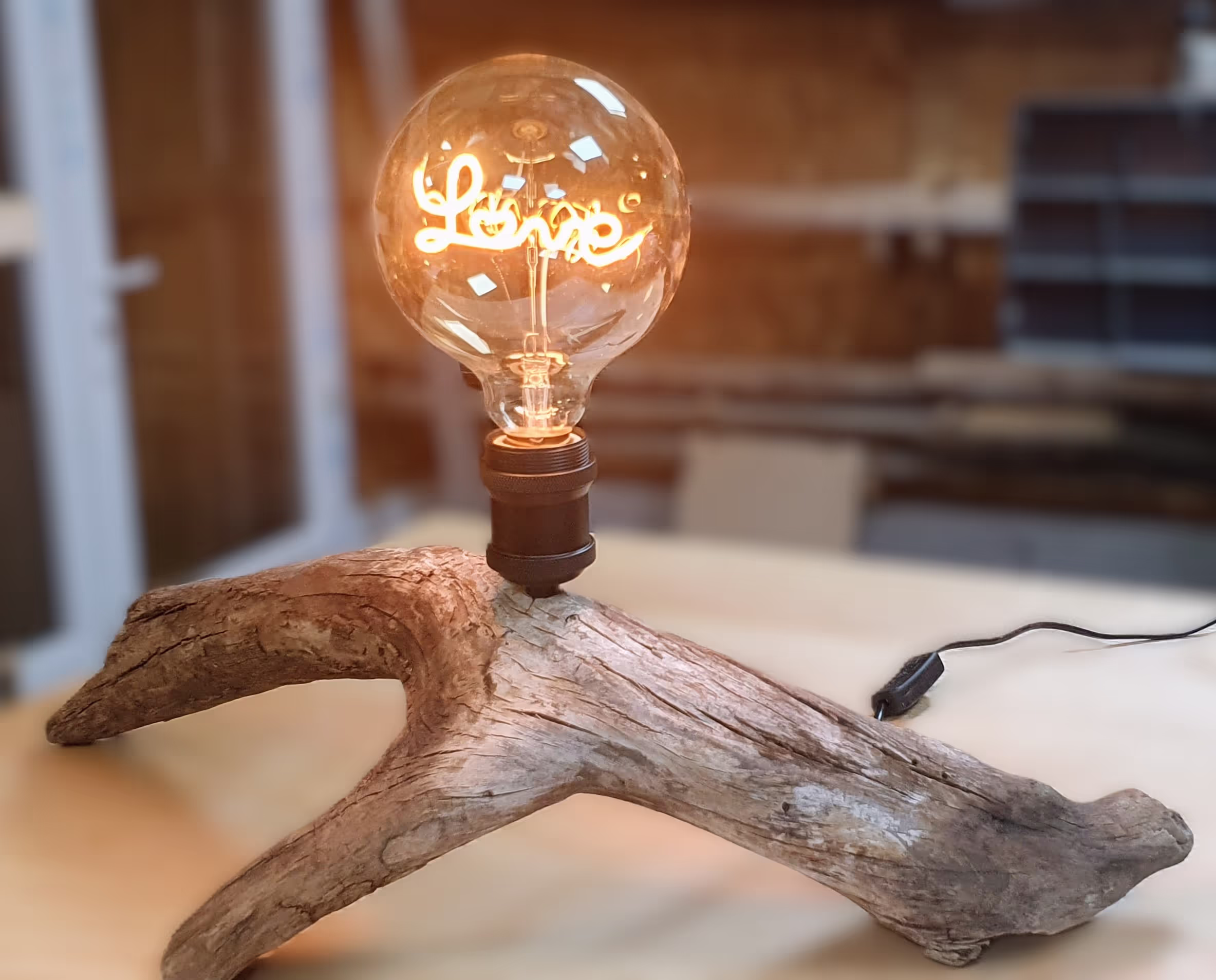 Lampe trépied en bois flotté avec ampoule à filament écrivant 'LOVE', photographiée dans mon atelier floutté avec un filtre sépia.