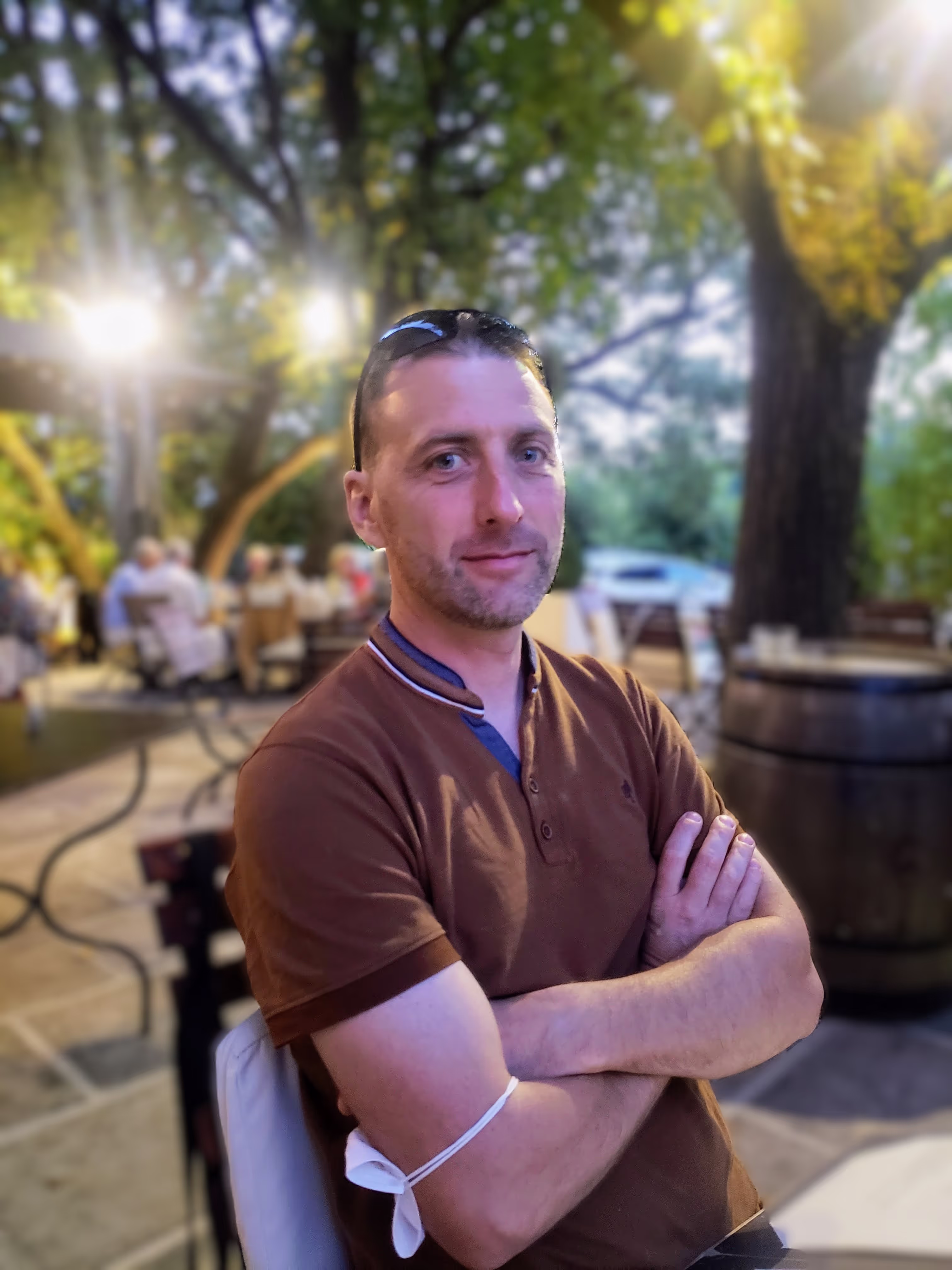 Thierry, artisan bois, assis à une terrasse, bras croisés sur le torse, portant un polo marron et des lunettes de soleil remontées sur la tête, regardant l'objectif de façon naturelle.