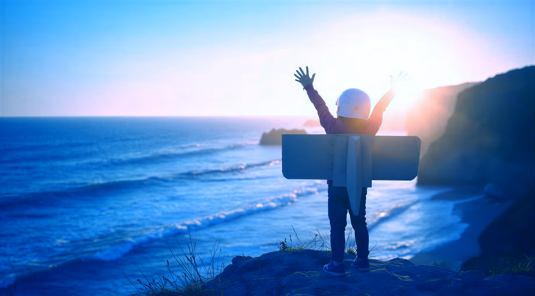 Kind mit Astronautenhelm und Spielzeugflugzeug steht am Klippenrand mit Blick aufs Meer bei Sonnenuntergang.