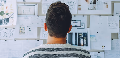 Person von hinten mit kurzen dunklen Haaren vor einer Wand mit vielen angehefteten Fotos und Dokumenten.