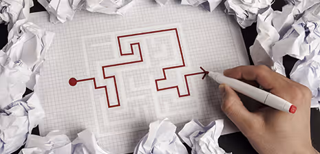 Hand draws a maze with red ink on graph paper surrounded by crumpled papers.