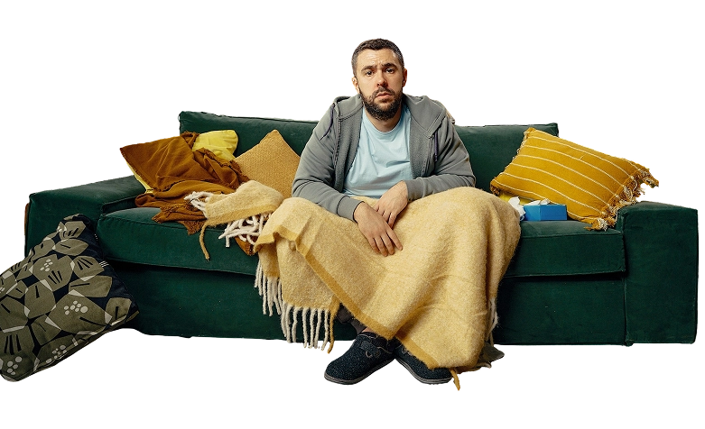 Imaginea prezintă un bărbat care are simptome de răceală și gripă, este acoperit cu o pătură galbenă și stă pe o canapea verde.
