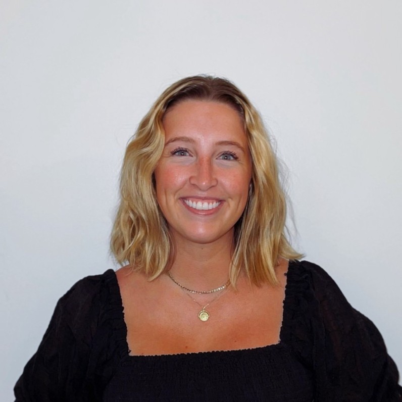 Smiling woman with shoulder-length blonde hair wearing a black top and a gold necklace against a plain white background.