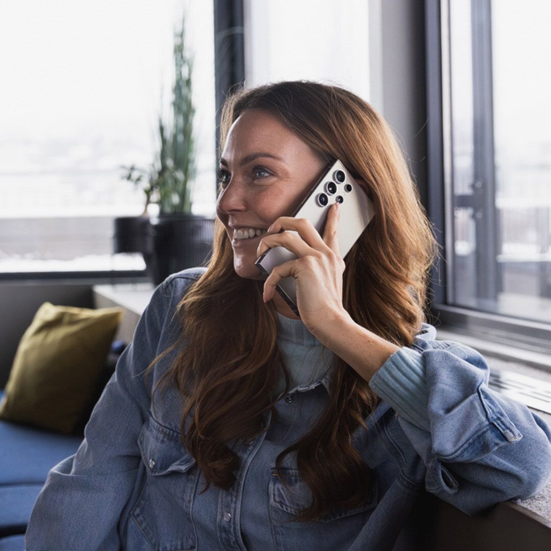 Smiling woman with long brown hair in denim jacket talking on a smartphone near a window.