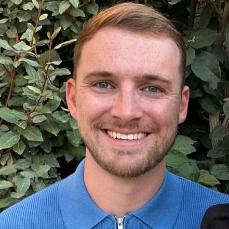 Smiling young man with short light brown hair and blue eyes wearing a blue zip-up sweater standing in front of leafy bushes.