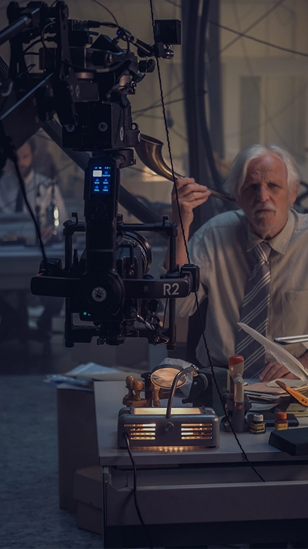 Ein älterer Mann mit weißem Bart sitzt verwirrt an einem Schreibtisch in einem dimmen, industriellen Raum von der Panda Pictures Filmproduktion München und Ulm und betrachtet Papiere unter einer alten Lampe, während ein Kamerastecker mit RED-Kamera-Rig im Vordergrund positioniert ist.