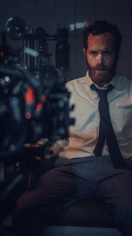 Ein bärtiger Mann mit einem Verband auf der Nase sitzt ernst in einem engen, beleuchteten Raum von der Panda Pictures Filmproduktion München und Ulm,, umgeben von industriellen Elementen wie Rohren und einer Kamera im Vordergrund, trägt ein weißes Hemd und graue Hose.