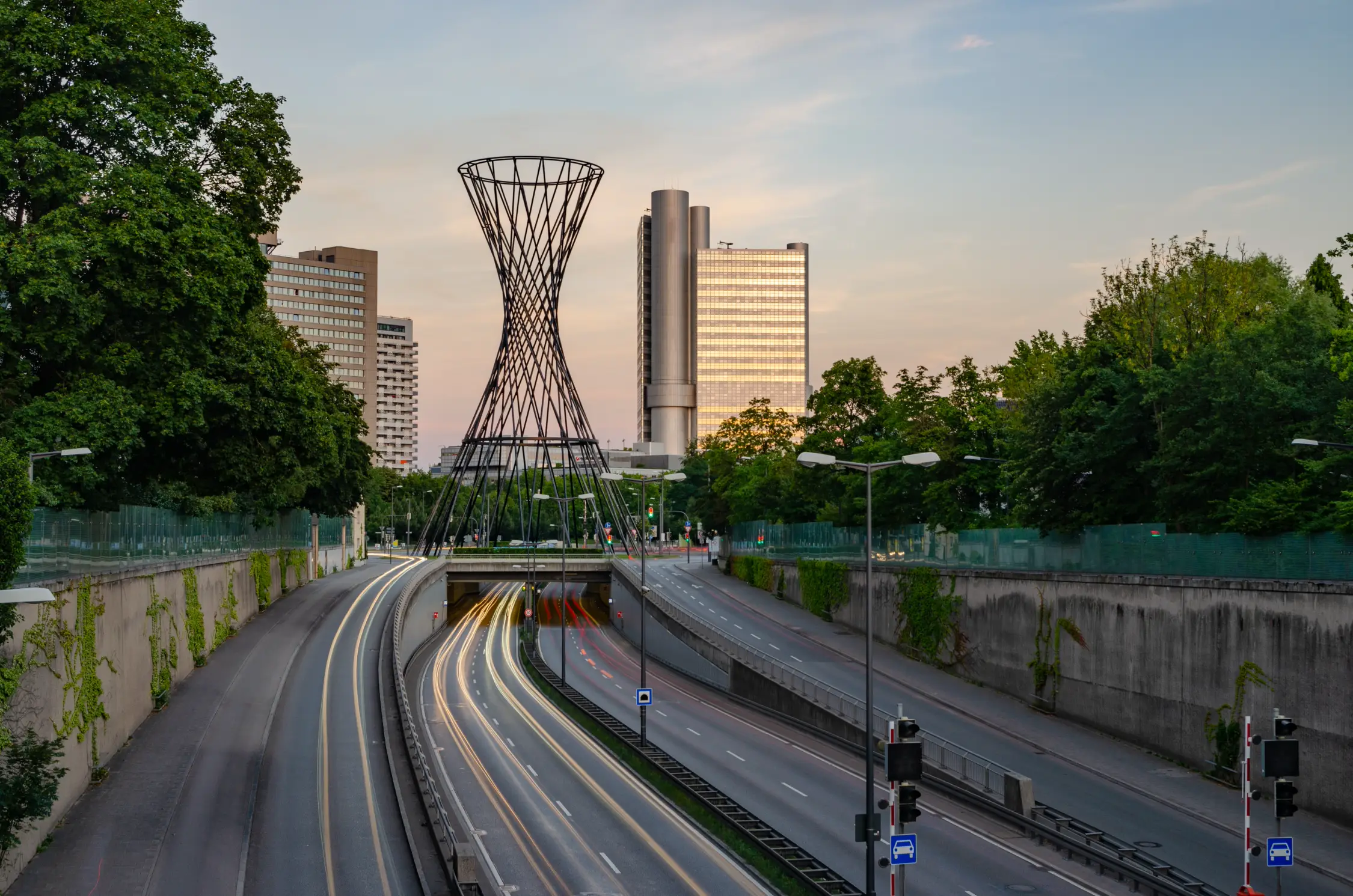Eine Dämmerungsaufnahme der Verkehrsanlage am Mittleren Ring in München mit der markanten skulpturalen, sich nach oben verjüngenden Metallstruktur in der Mitte. Autos hinterlassen auf der Straße Lichtspuren, eingerahmt von Bäumen und modernen Hochhäusern – eine städtische Aufnahme, die die Standorte der Panda Pictures Filmproduktion München Ulm Videoproduktion verbindet.
