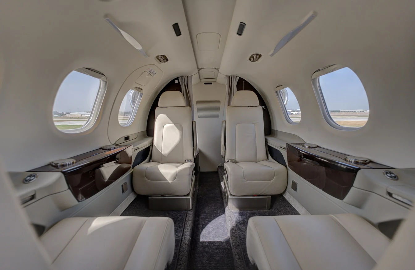 Interior view of a private jet cabin with four cream leather passenger seats and large windows.