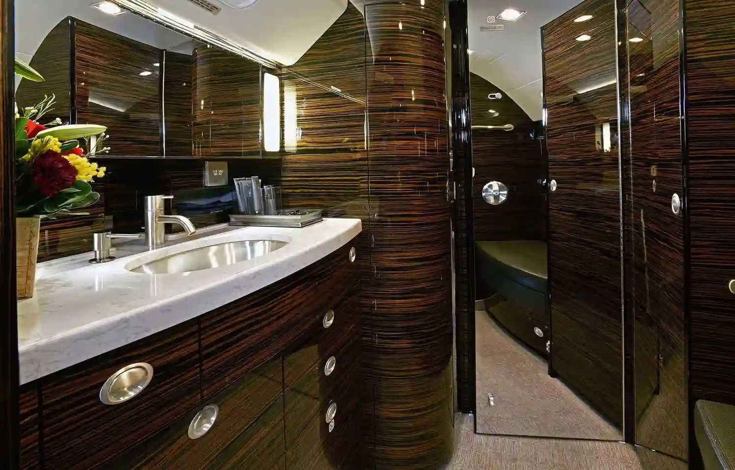 Luxurious private jet lavatory with marble counter, sink, and dark wood veneer cabinets.