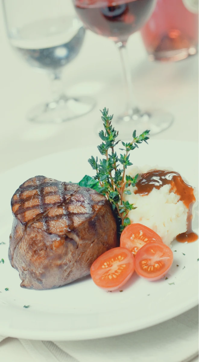 Plated meal of grilled steak, mashed potatoes, and tomatoes, with wine glasses in the background.