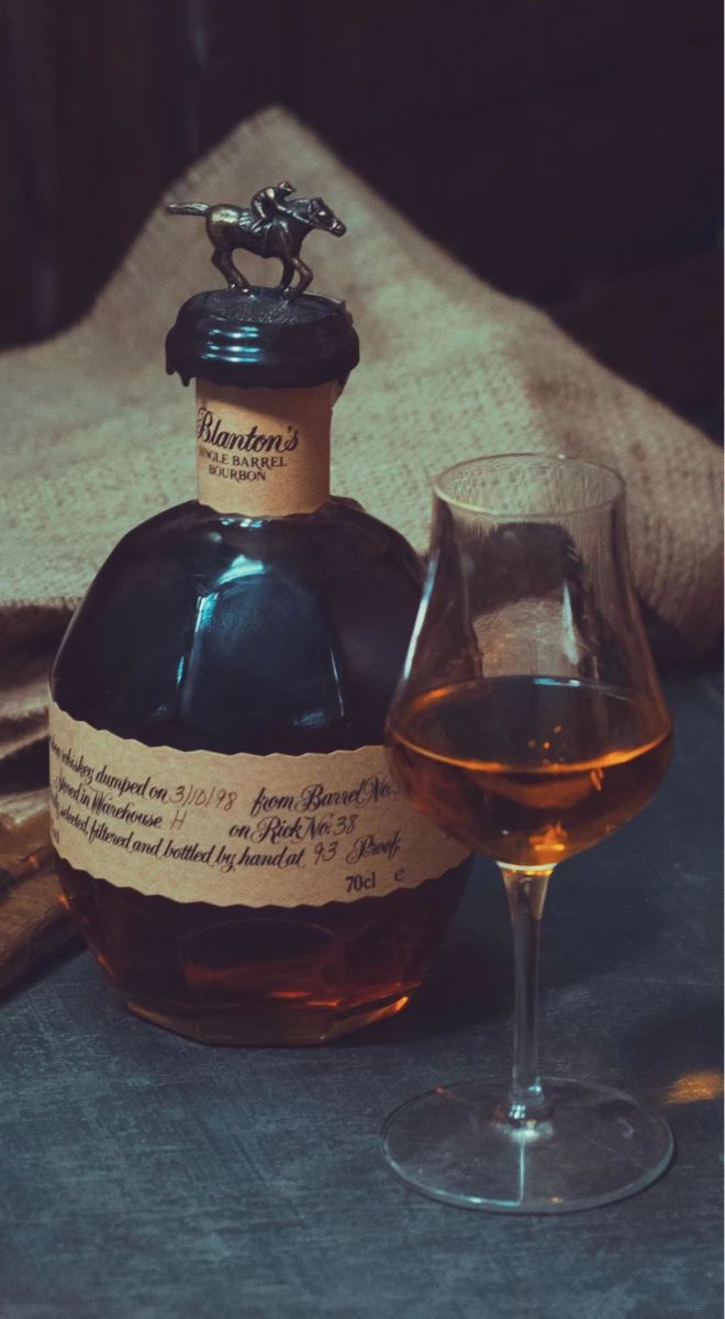 Still life of a bottle of Blanton's Single Barrel Bourbon with a horse stopper next to a glass of whiskey.