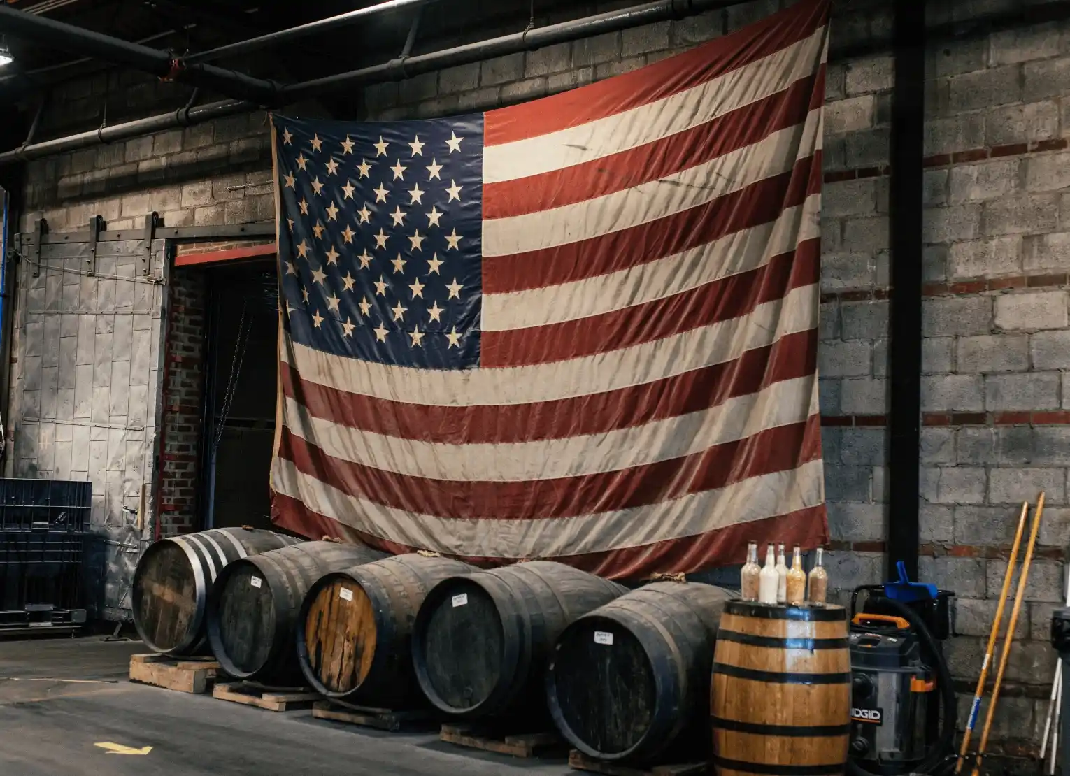 Large American flag hanging over wooden whiskey barrels in a dimly lit industrial space.