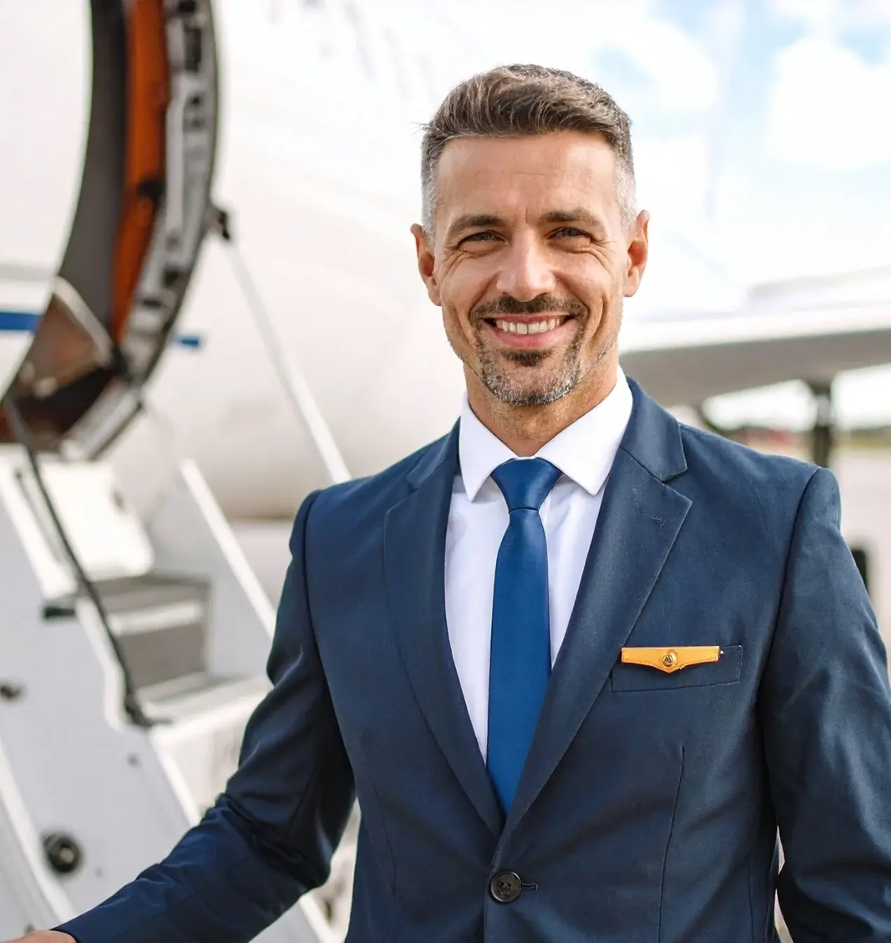 Smiling man in a blue suit and tie standing by the open door of a private jet.