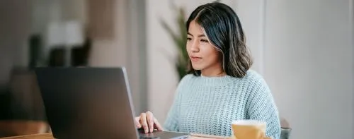 A young woman wearing a light blue sweater works on her laptop with focus. A coffee cup is placed on the table beside her in a cozy indoor setting