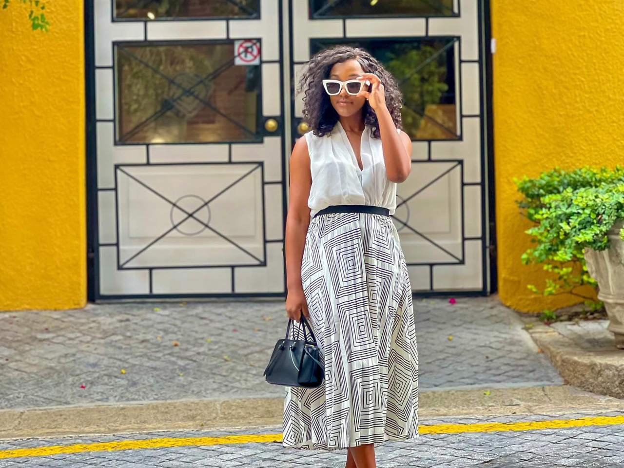 Woman in a white blouse and geometric-patterned skirt holding a black purse and adjusting sunglasses.