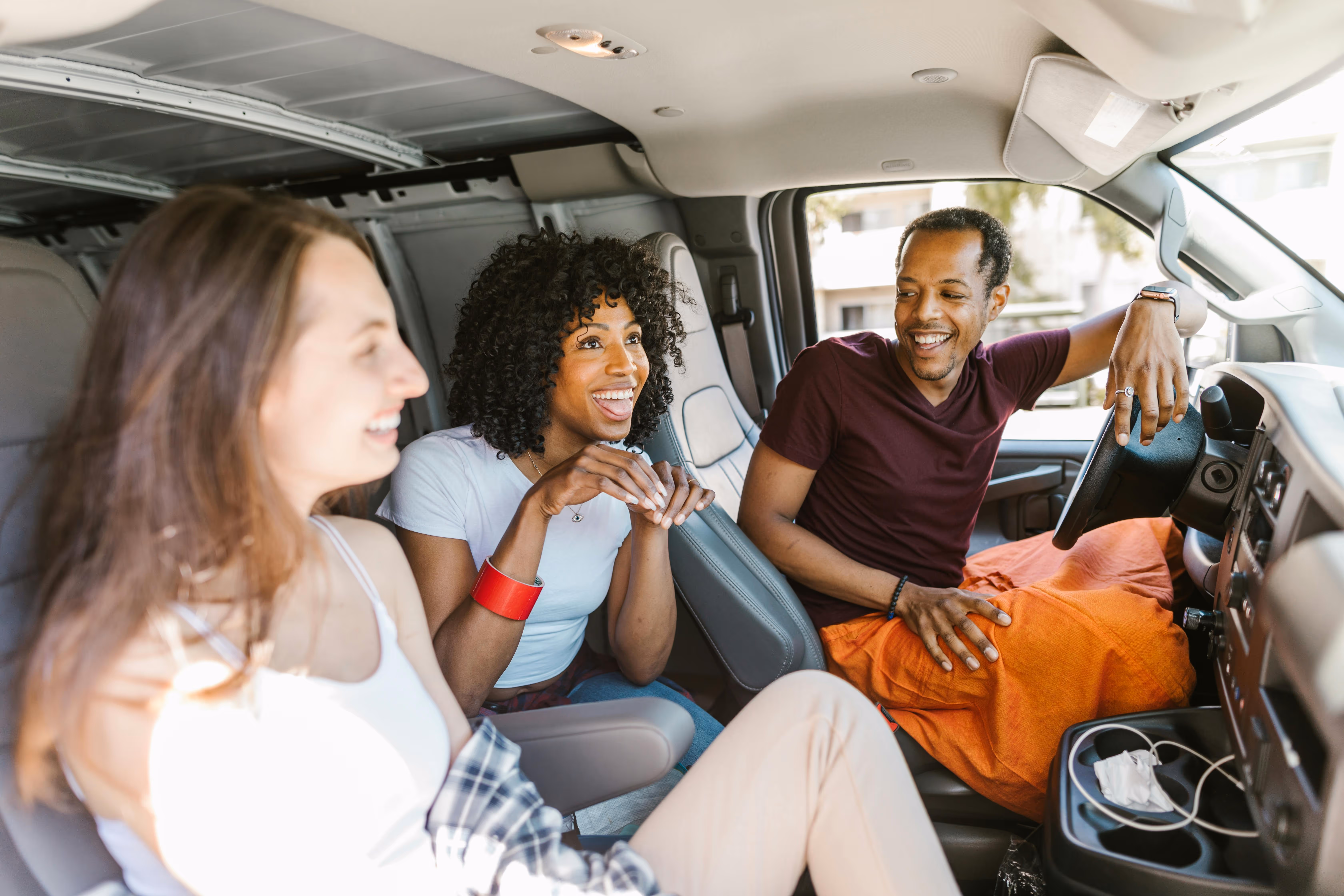 Three friends sitting and smiling inside a parked vehicle, two women and one man in front seats.
