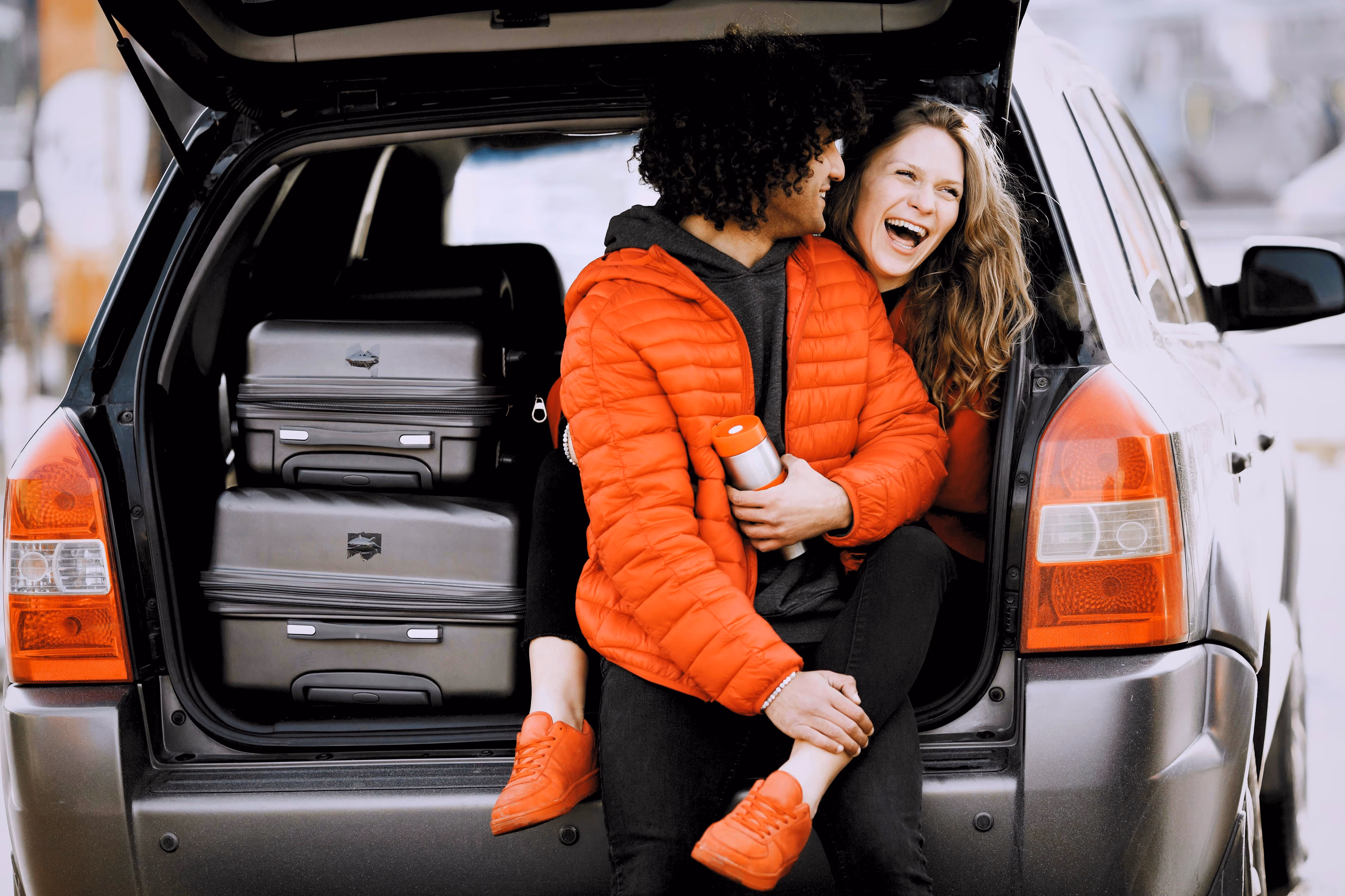Couple sitting in the open trunk of a car with packed suitcases, laughing and wearing red jackets.