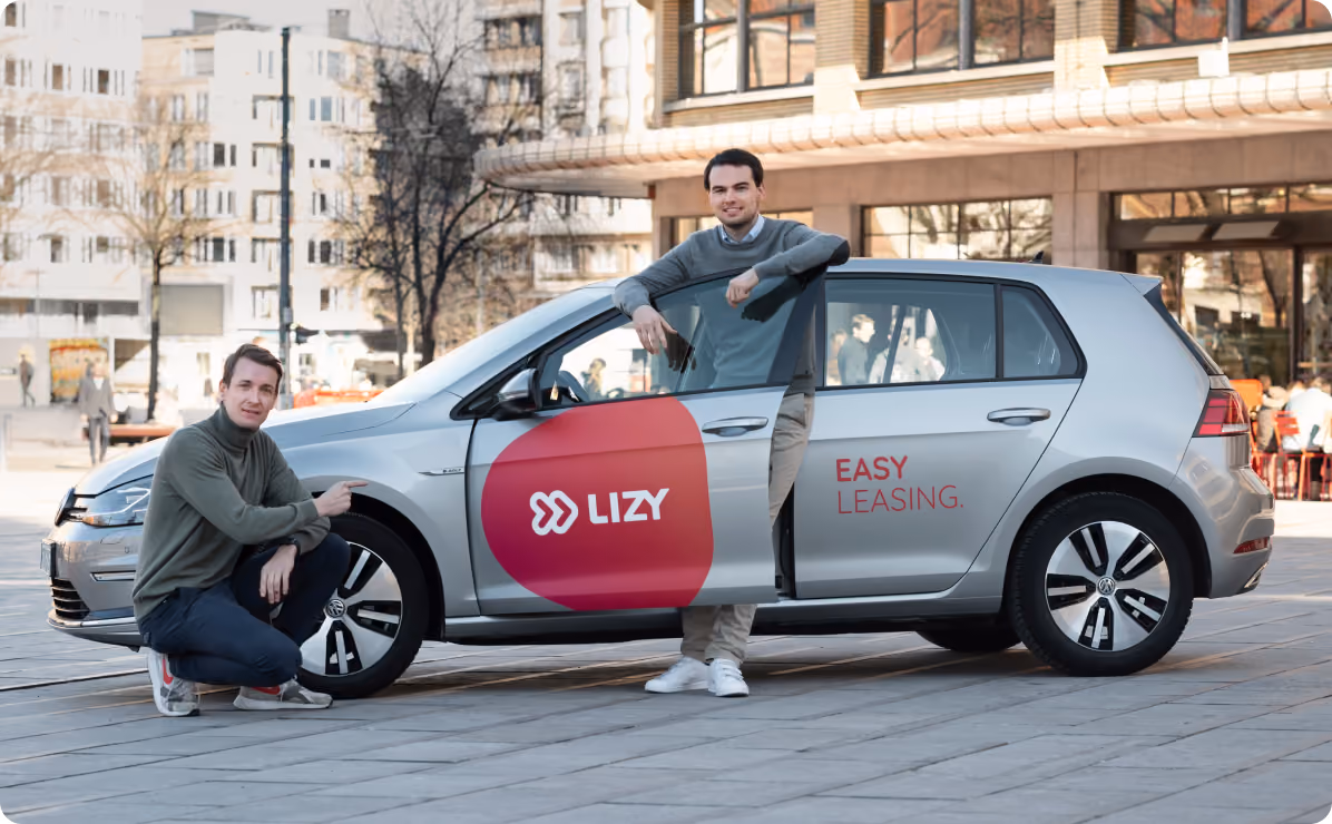 Twee mannen poseren bij een grijze auto met de tekst 'LIZY' en 'EASY LEASING' in een stedelijke omgeving.