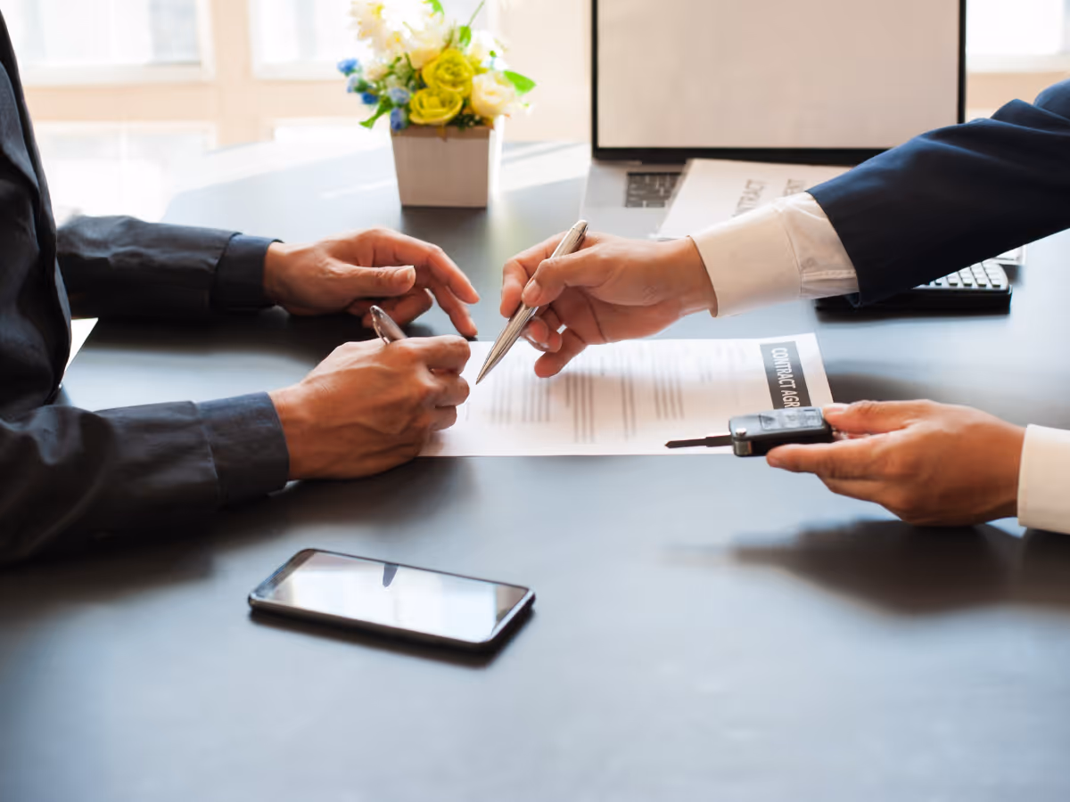 Twee personen ondertekenen een contract aan een bureau met een autosleutel en een smartphone erop.