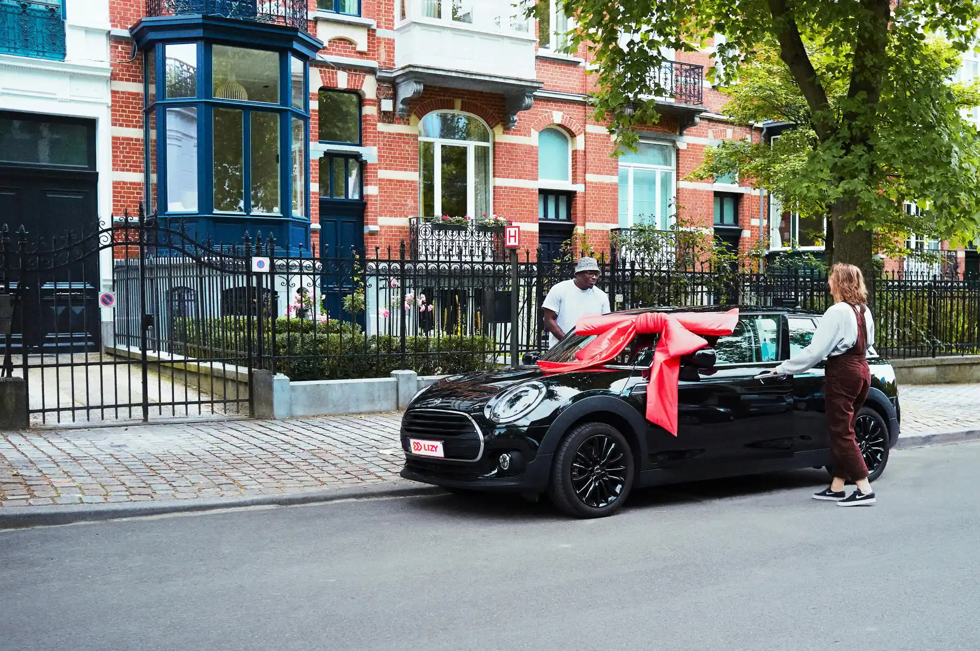 Black Mini Cooper car with a large red bow on the hood parked on a street in front of brick townhouses with two people nearby.