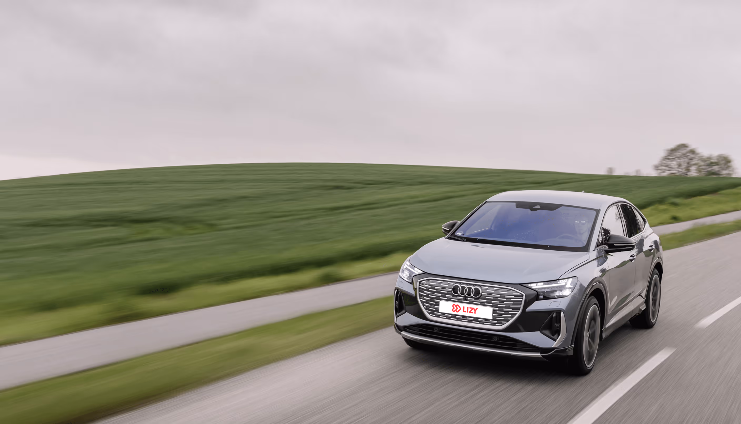 Gray Audi electric SUV driving on a paved road beside green fields under an overcast sky.