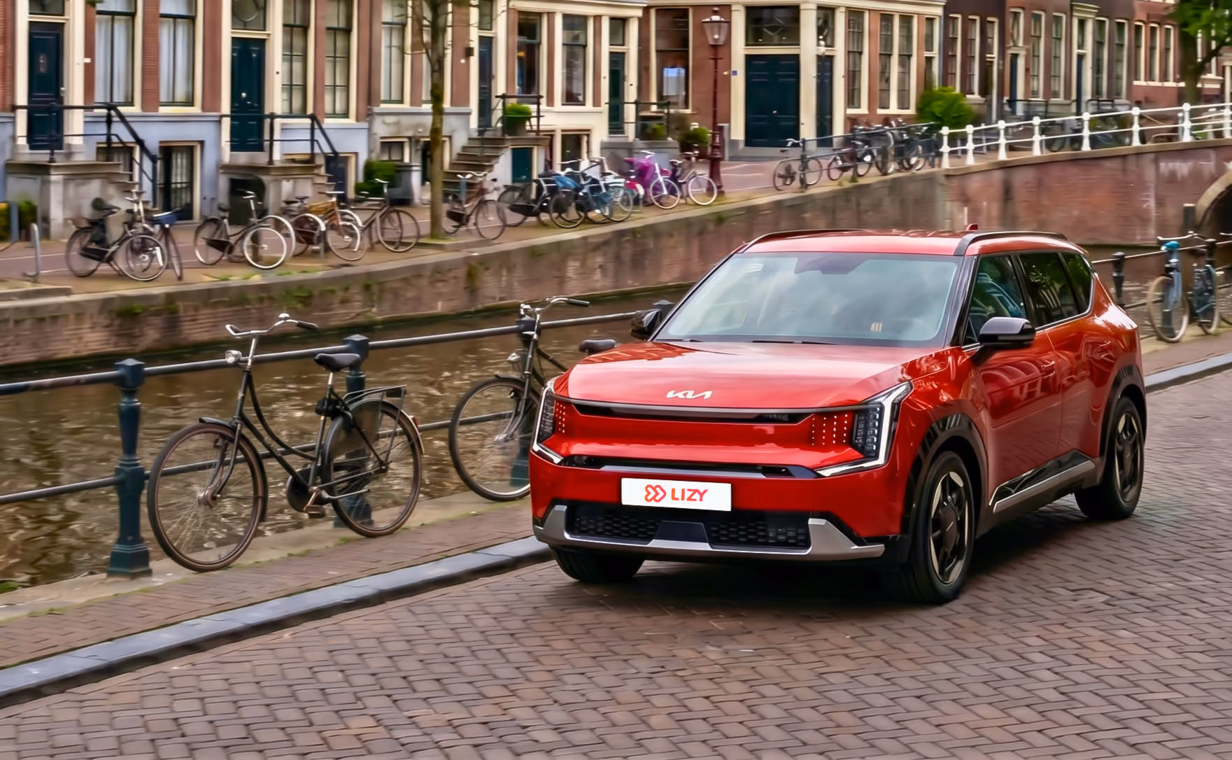 Een jong gebruikte rode KIA EV3 van LIZY geparkeerd langs een gracht in Amsterdam met fietsen en grachtenpanden op de achtergrond.