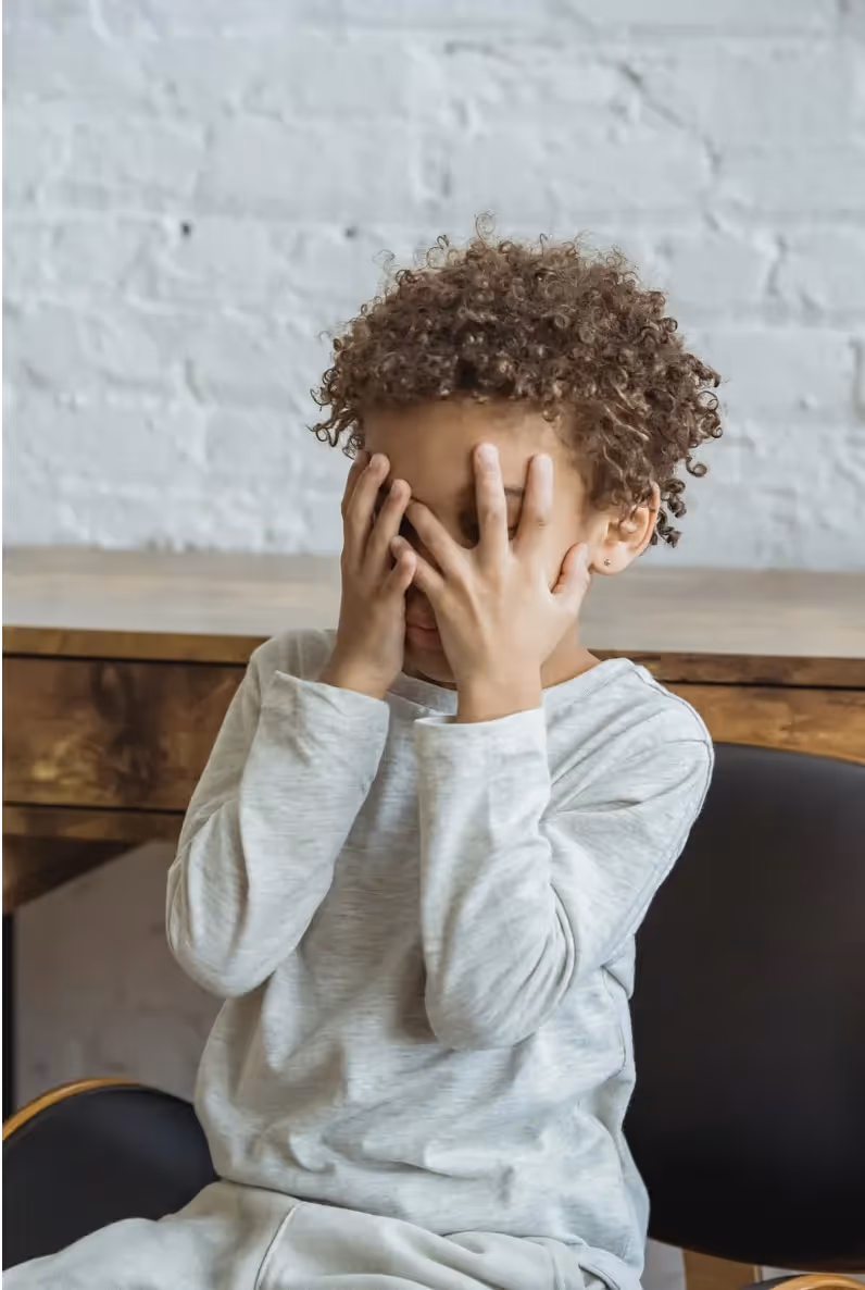A child sitting in a chair covering their face with their hands
