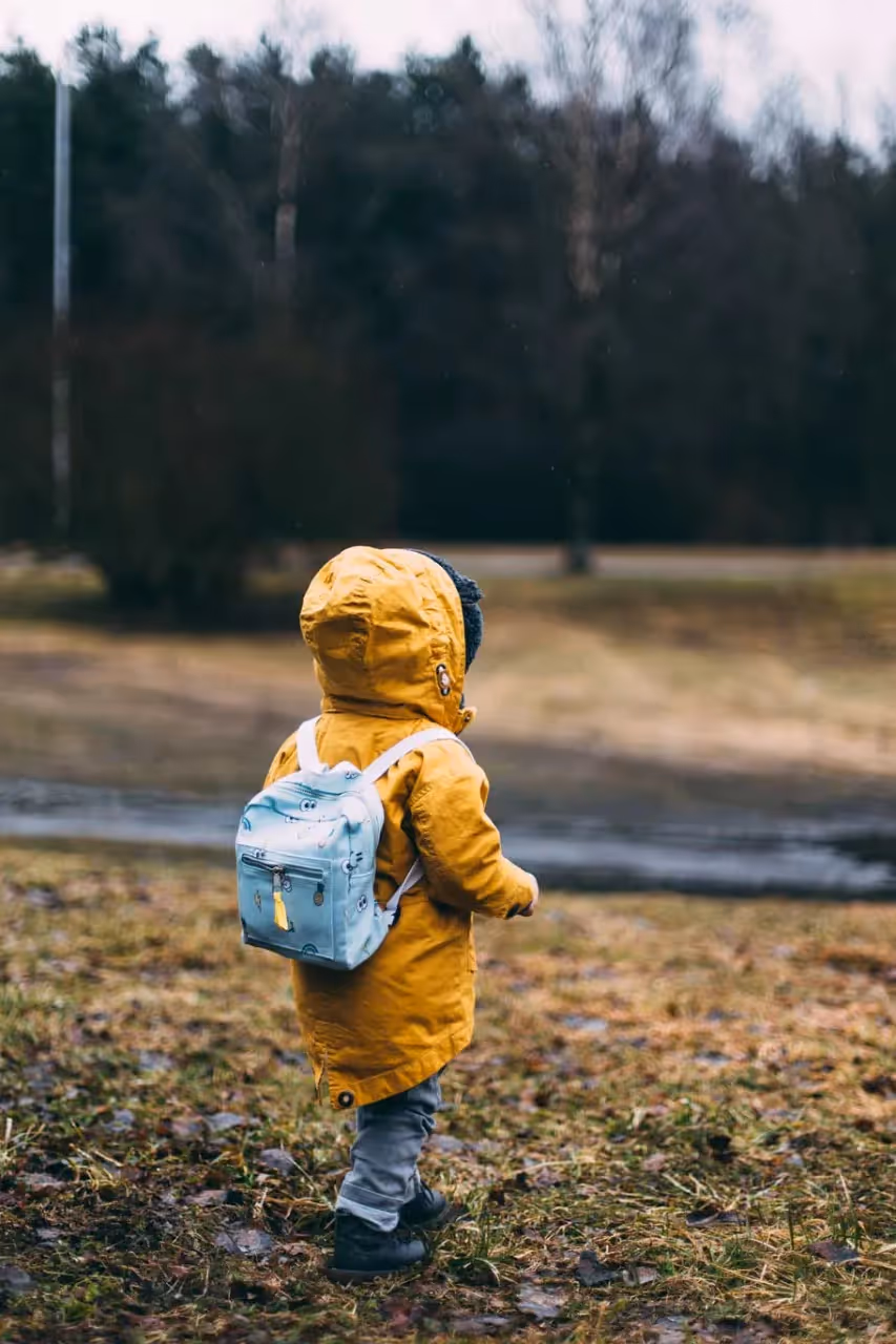 Child wearing a yellow jacket and blue backpack walking on grass.