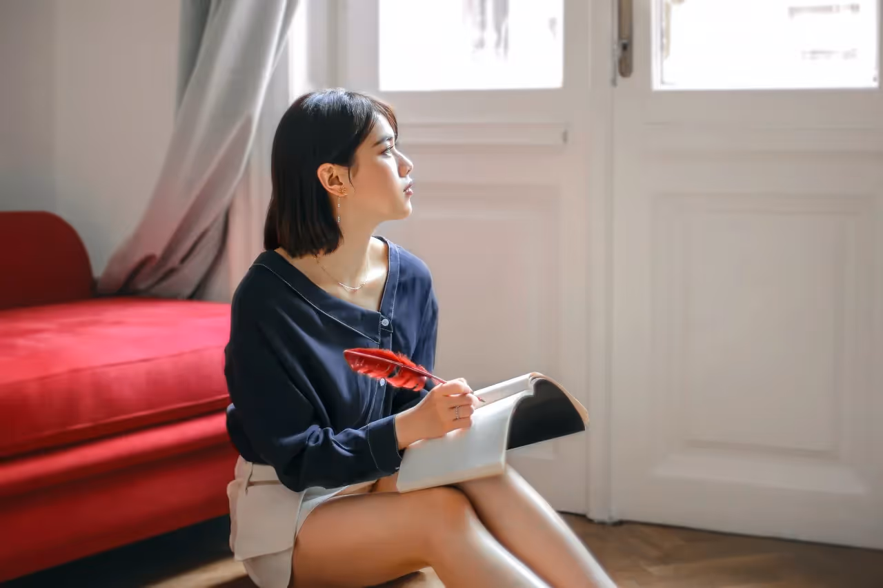 a woman sitting on the floor writing in a journal with a red feather pen